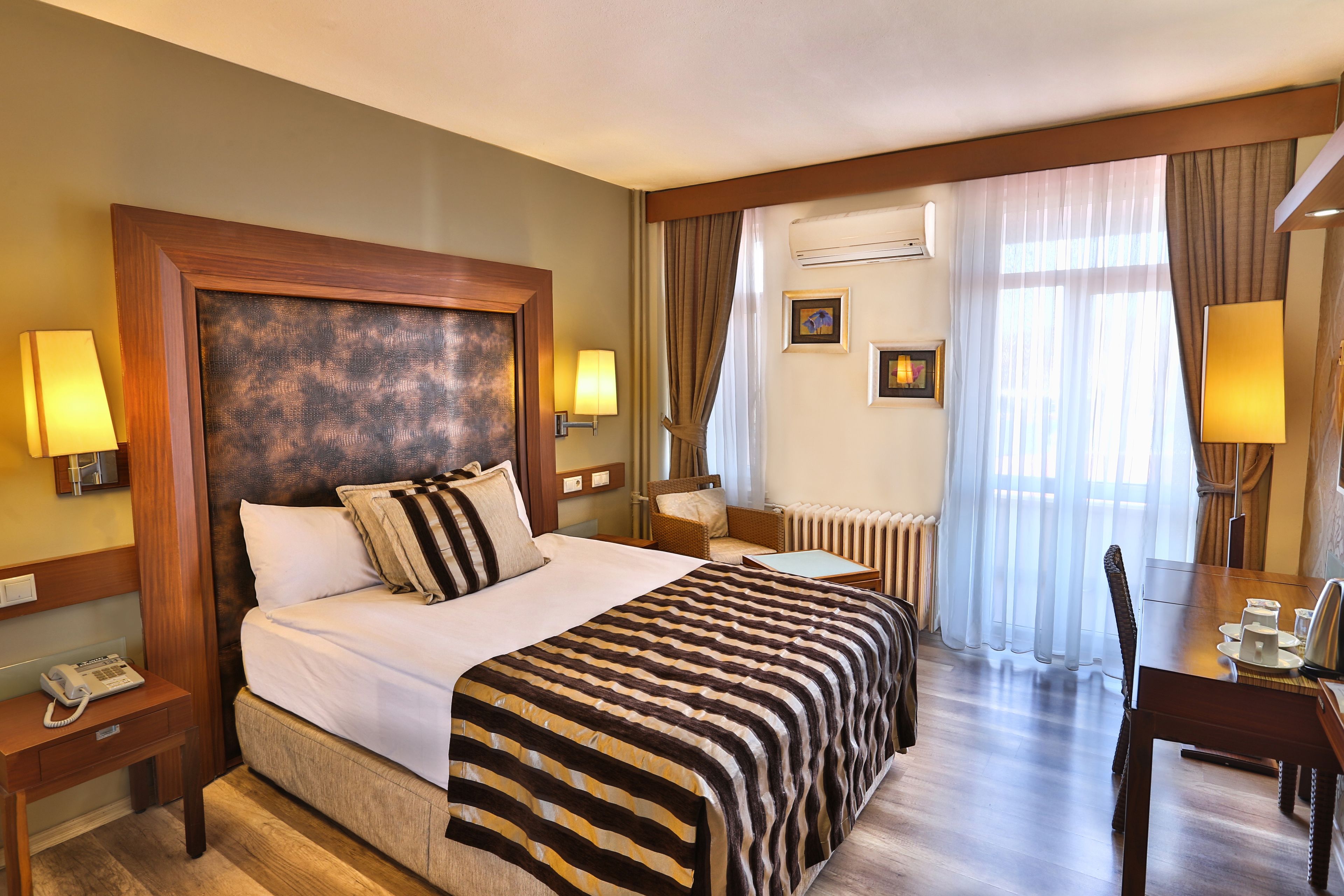 A hotel room with a large bed, two bedside tables with lamps, a chair, a small table, and a view of the outside through sheer curtains.