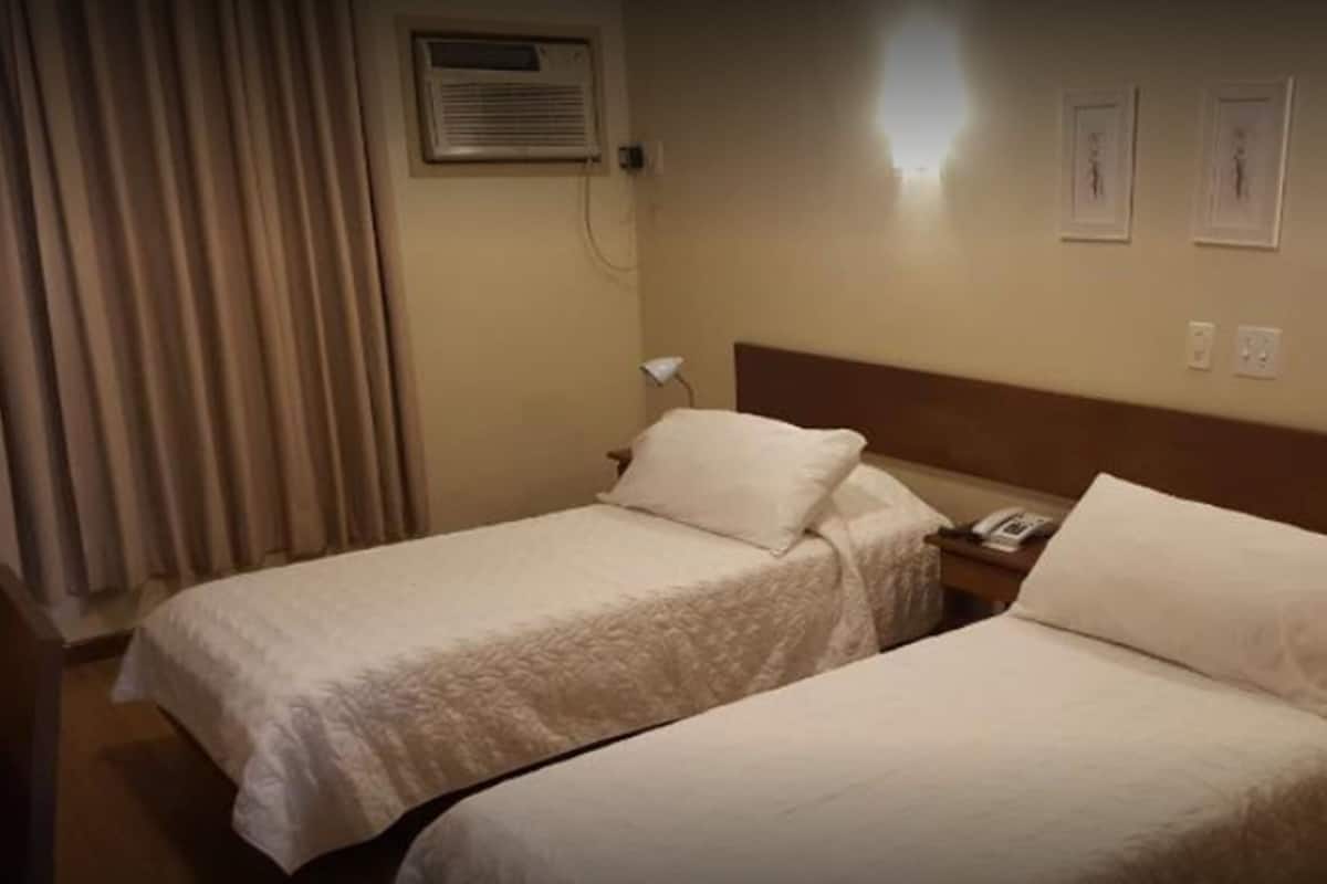 A hotel room with two single beds, a bedside table with a lamp, an air conditioning unit, and two framed pictures on the wall.