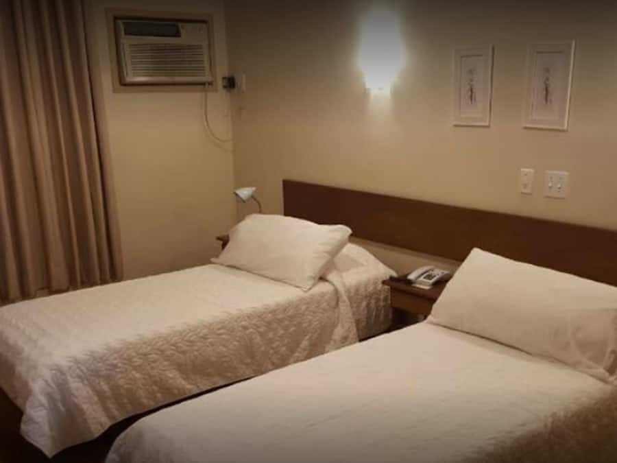 Habitación de hotel con dos camas individuales, una mesita de noche con lámpara, un equipo de aire acondicionado y dos cuadros en la pared.