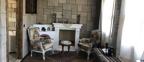 A cozy room with stone walls, a fireplace, two ornate chairs, a small table, and a patterned rug.