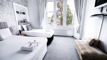 A hotel room with a large bed, a smaller bed, a sofa, a TV, and a window with a view of trees and buildings.