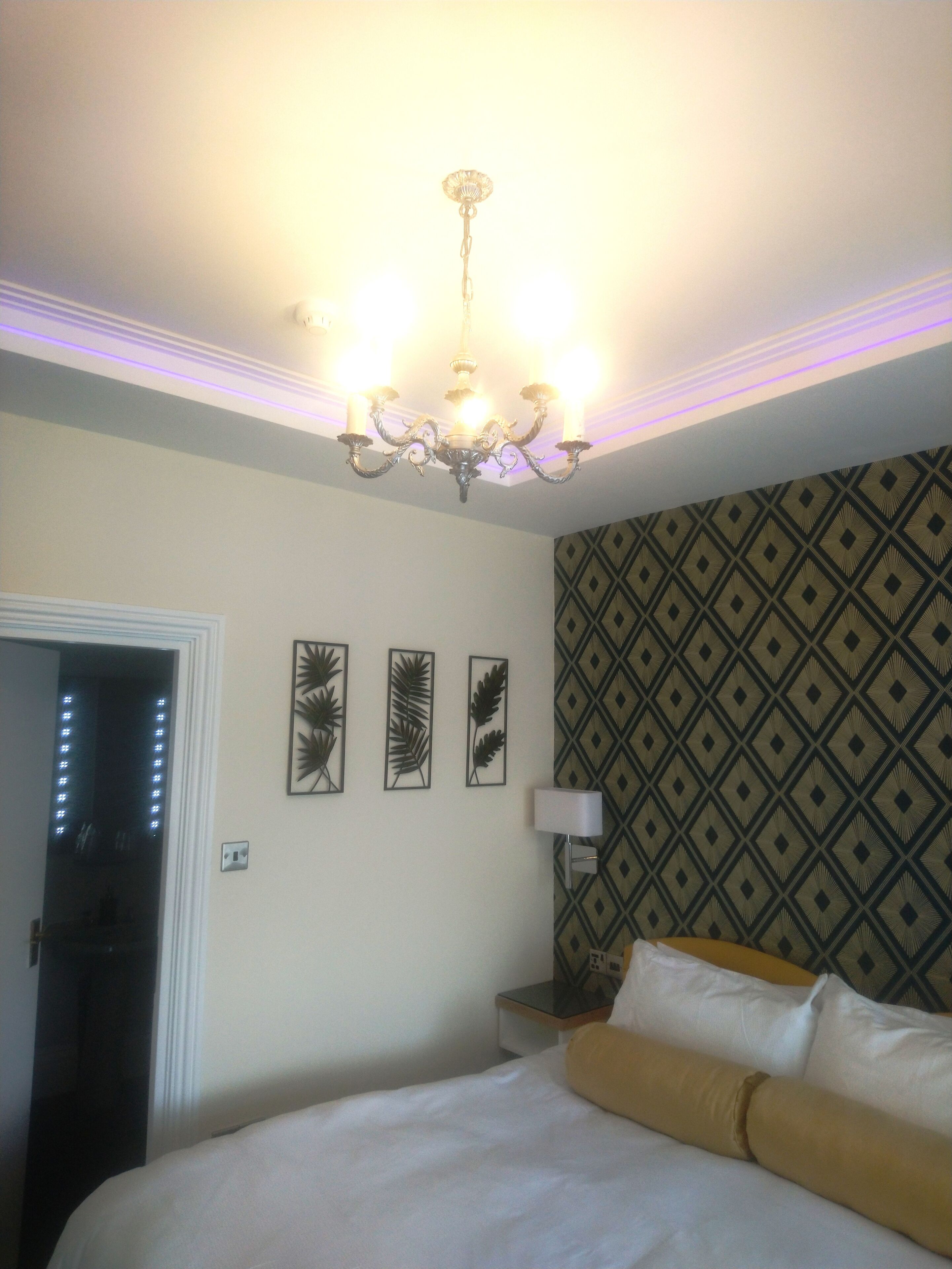 A hotel room with a bed, a chandelier, a wall with a geometric pattern, and three framed leaf decorations.