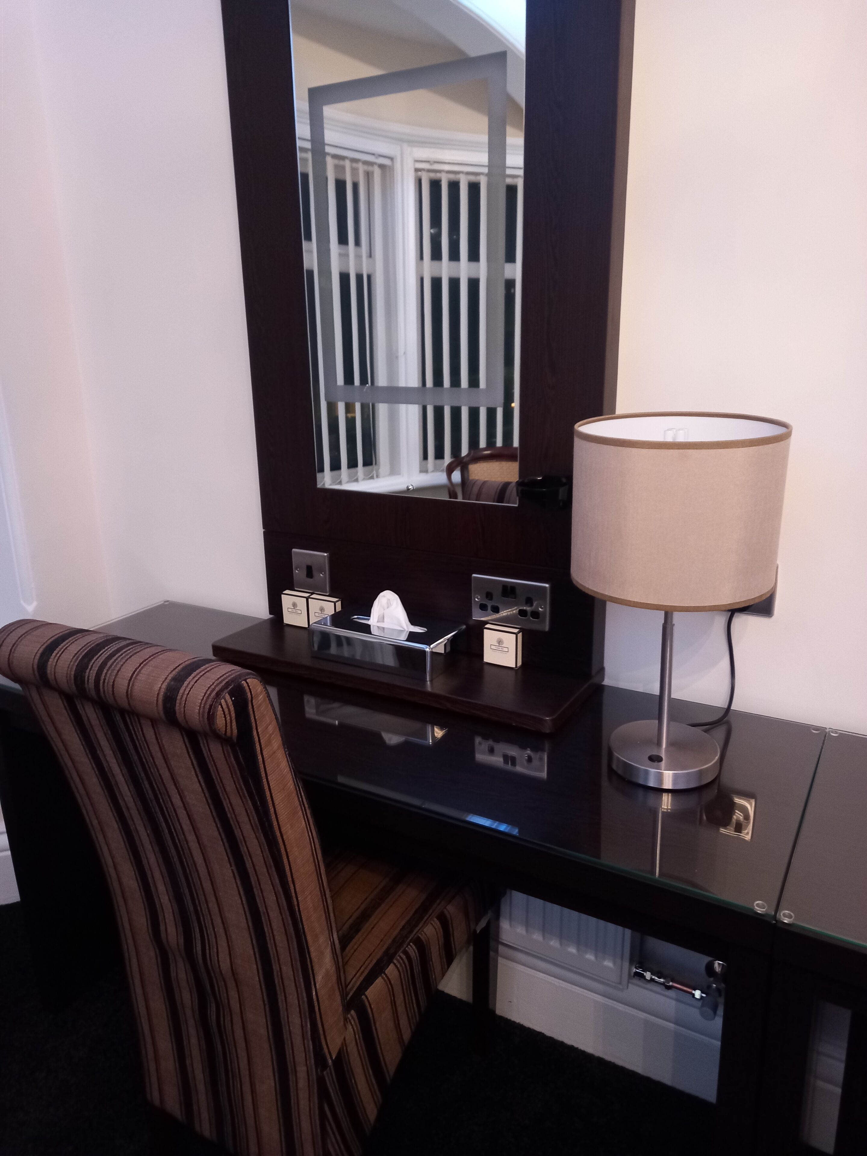 Ein Hotelzimmer mit dunklem Holztisch, einem Streifenstuhl, einem Spiegel, einer Lampe und einer kleinen Taschentuchbox.