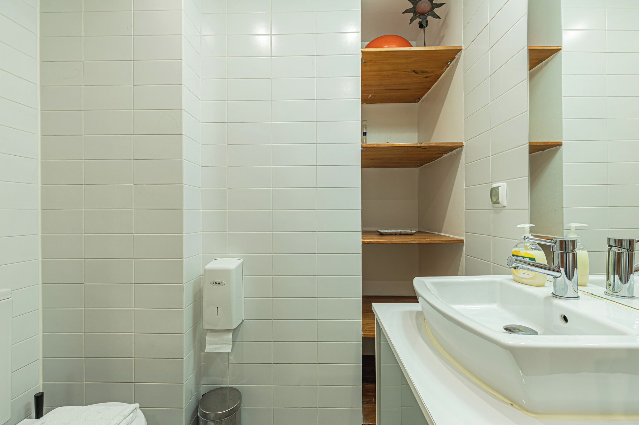A bathroom with white tiled walls, a white sink with chrome fixtures, a soap dispenser, and a trash can.