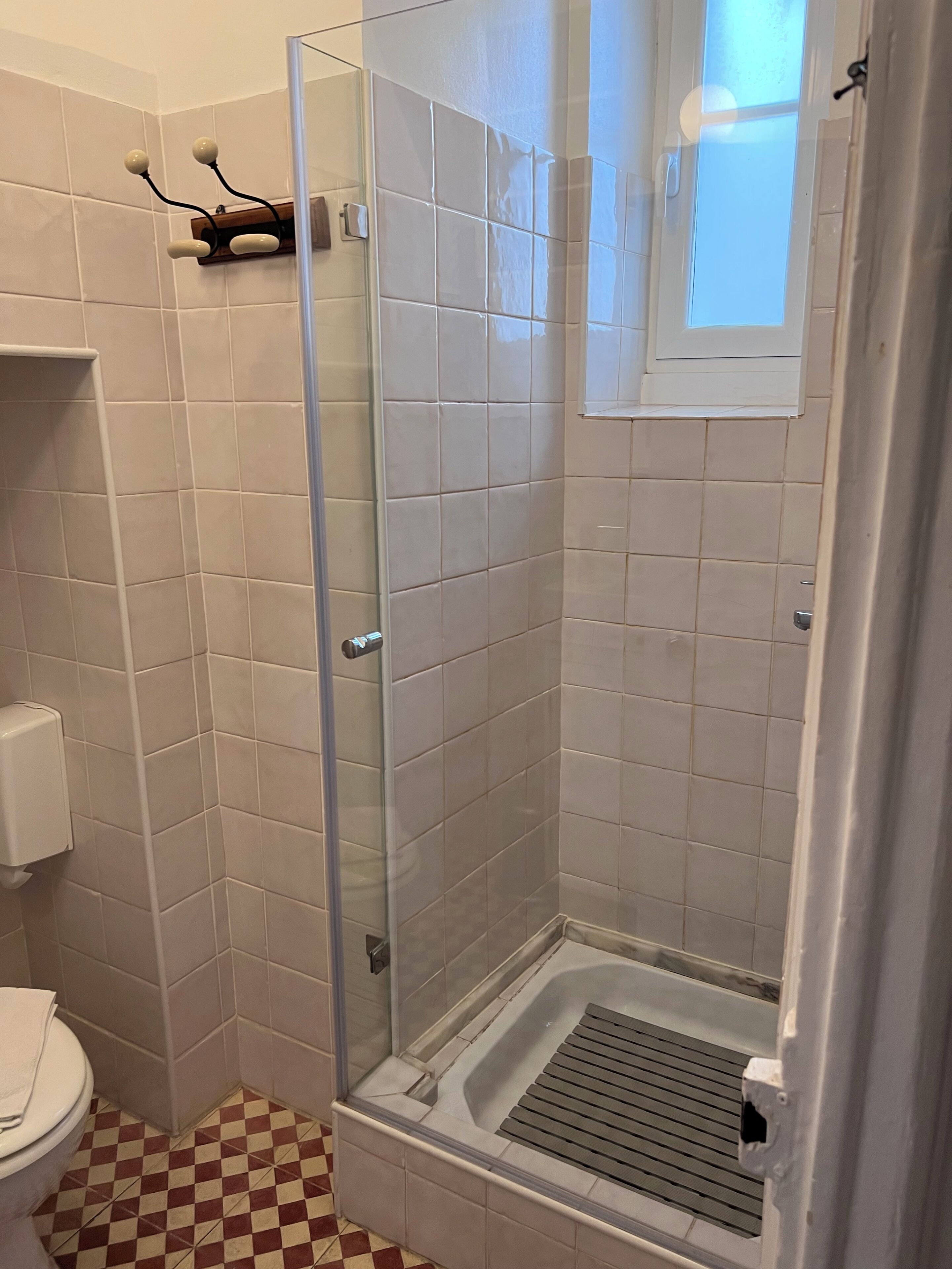 Un baño con cabina de ducha de vidrio, paredes cubiertas de azulejos blancos, suelo de cuadros y una ventana sobre la ducha.