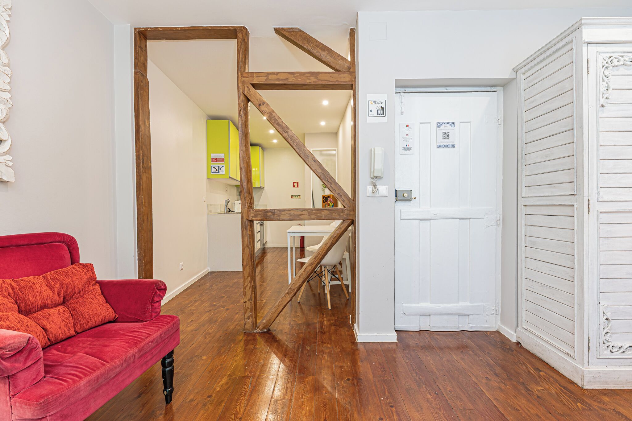 Quarto com sofá vermelho, vigas de madeira, porta branca e cozinha ao fundo.