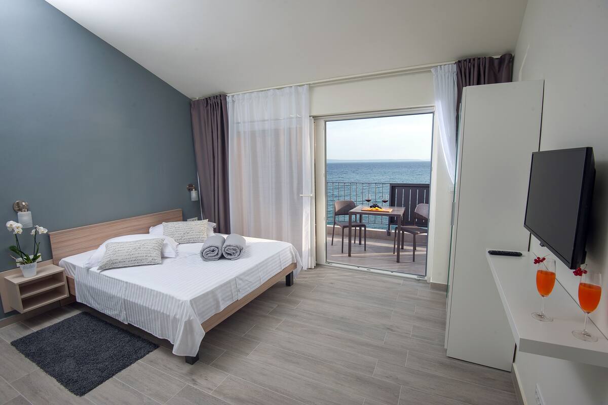 A hotel room with a bed, a desk with a TV, and a balcony with a view of the sea.