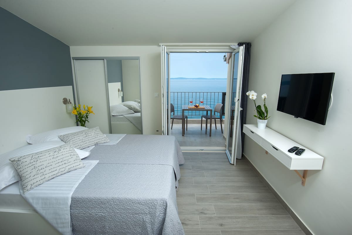 A hotel room with two beds, a balcony with a table and chairs, a TV mounted on the wall, and a small plant on a console table.