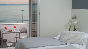 Un dormitorio con cama, una mesita con un plato y dos bebidas, una silla y un balcón con vista al mar.