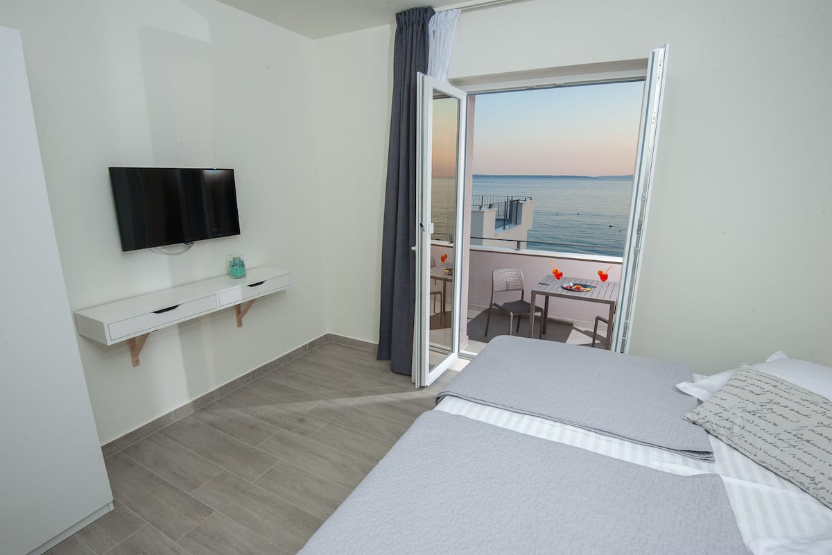 A hotel room with a bed, a TV, a small desk with a chair, and a balcony with a view of the sea.