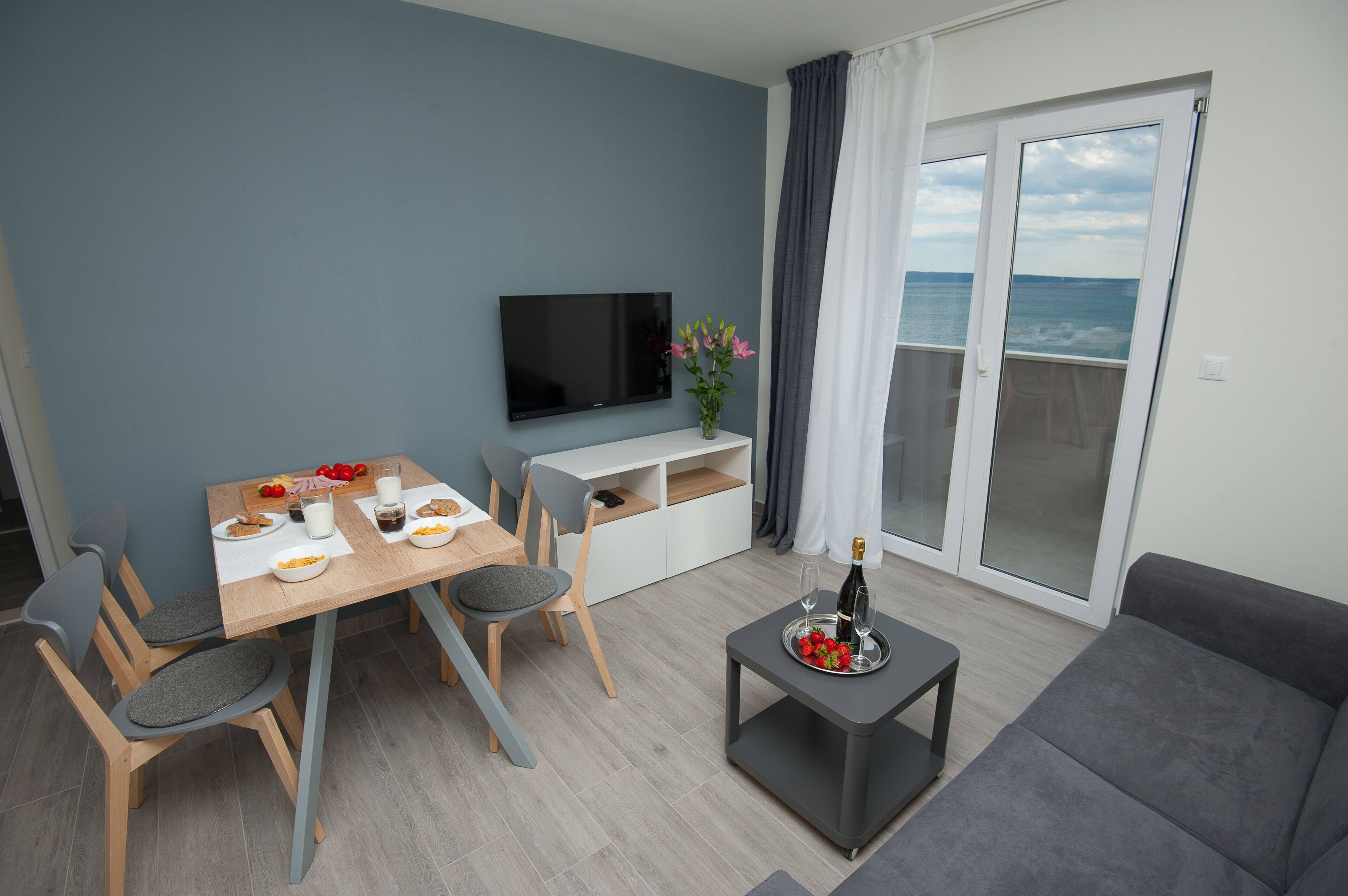 A modern living room with a dining table, chairs, a sofa, a TV, and a balcony with a view of the sea.