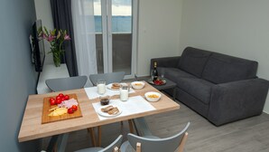 Un comedor con una mesa de madera preparada para el desayuno, un sofá gris y vista al mar a través de un ventanal.
