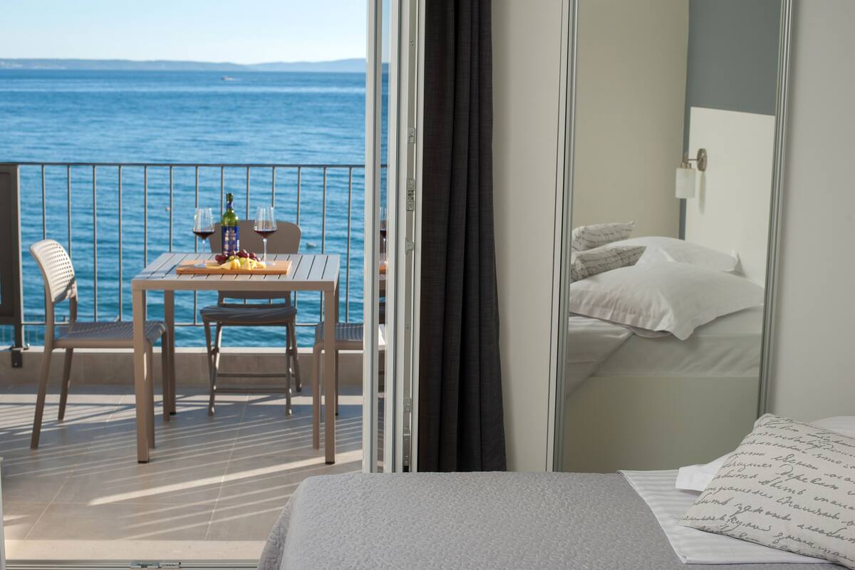 A balcony with a table set for two, a bed with white linens, and a view of the sea.