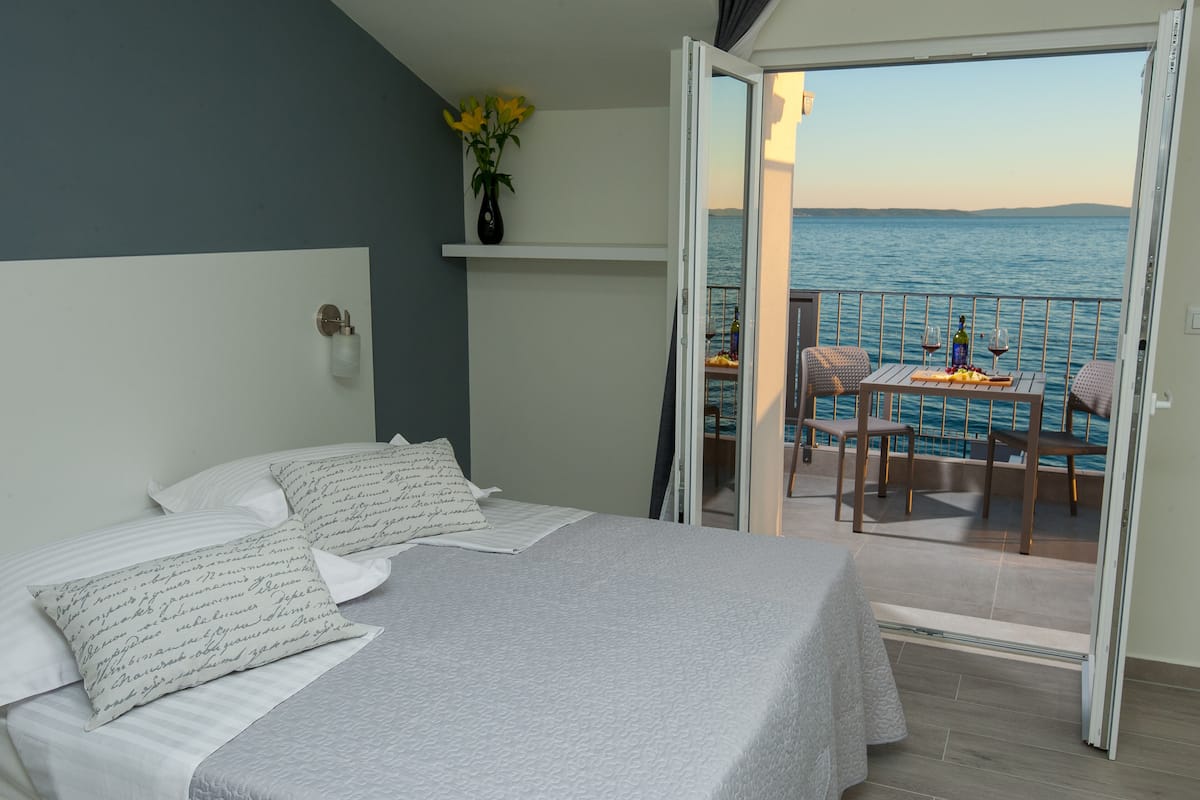 A bedroom with a bed, pillows, a nightstand, a vase with flowers, and a balcony with a table, chairs, and a view of the sea.