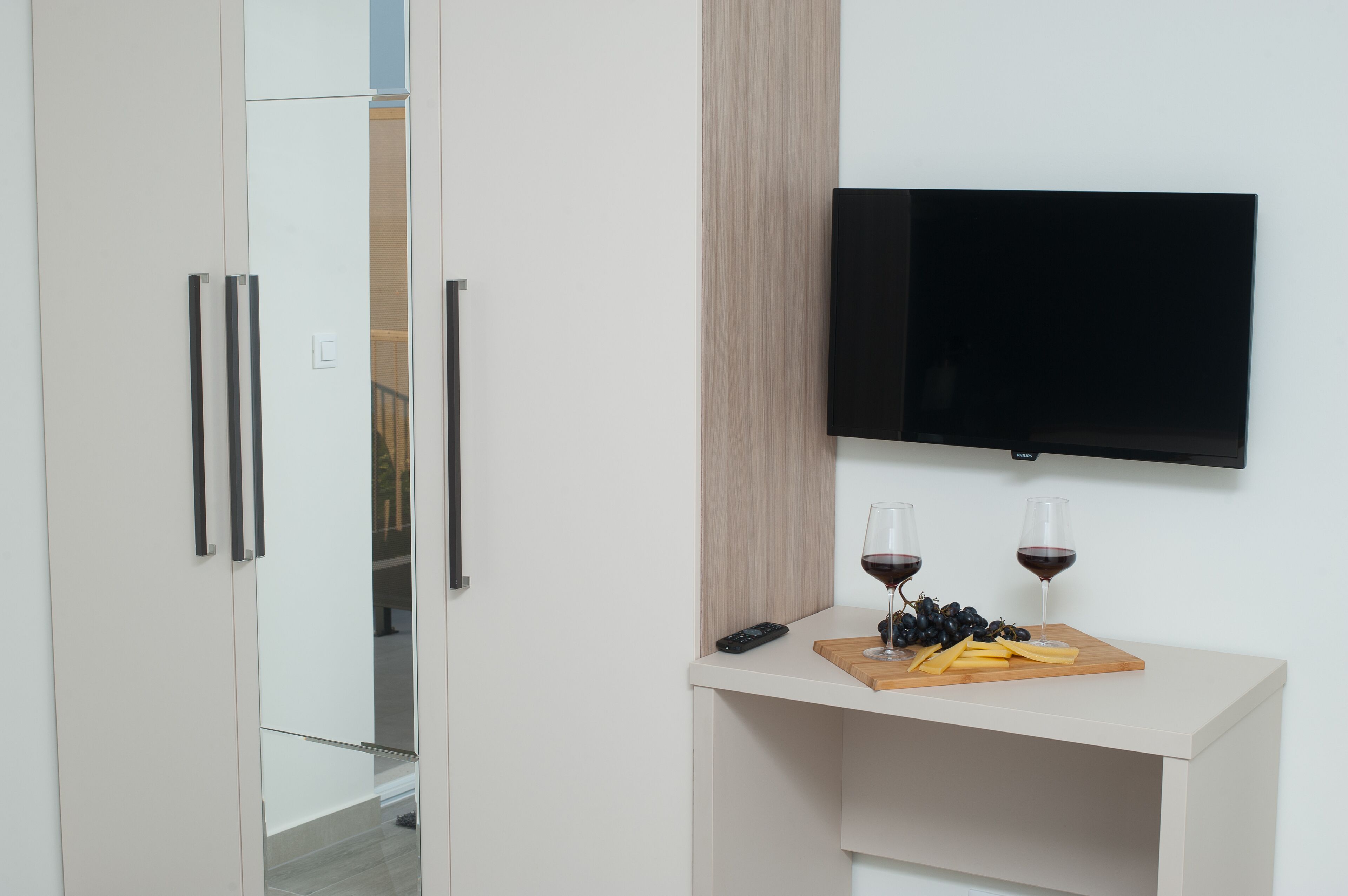 Ein modernes Zimmer mit an der Wand montiertem Fernseher, einem Holztisch mit Trauben und Käse sowie einer Fernbedienung auf dem Tisch.