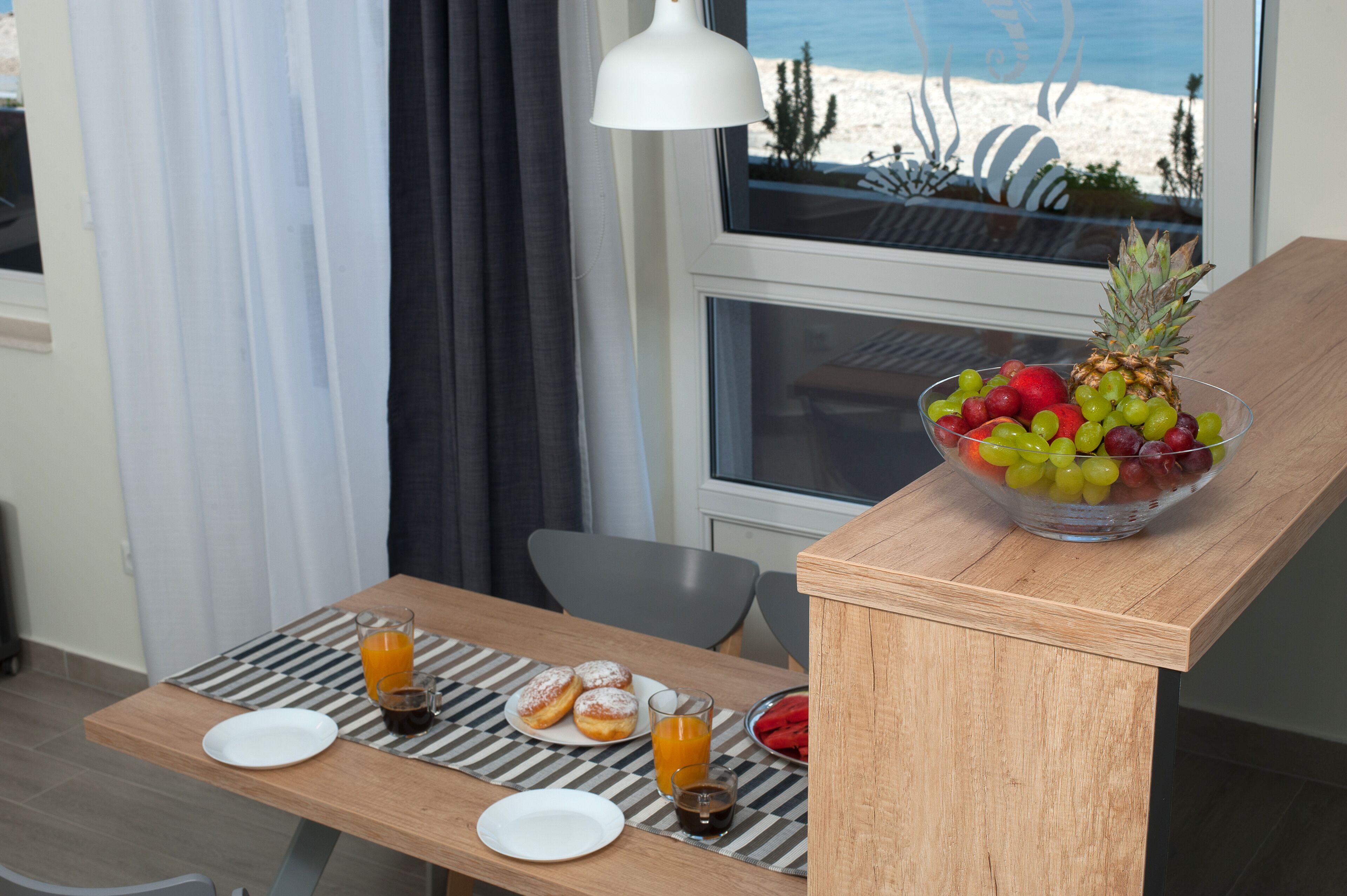 A breakfast setup with a fruit bowl, juice, and pastries on a wooden table with a view of the beach.