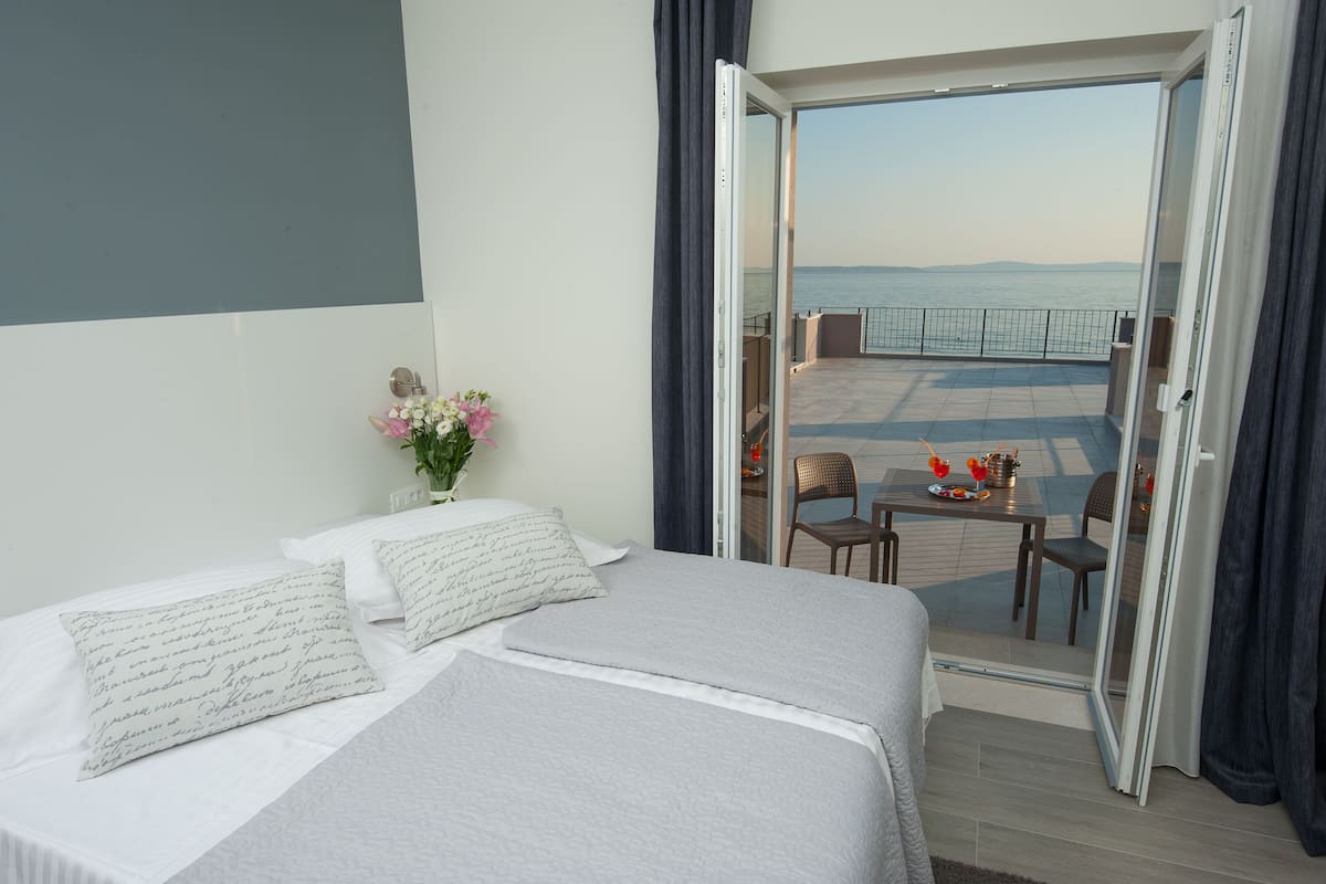 A hotel room with a bed, a small table with a chair, and a balcony with a view of the sea.