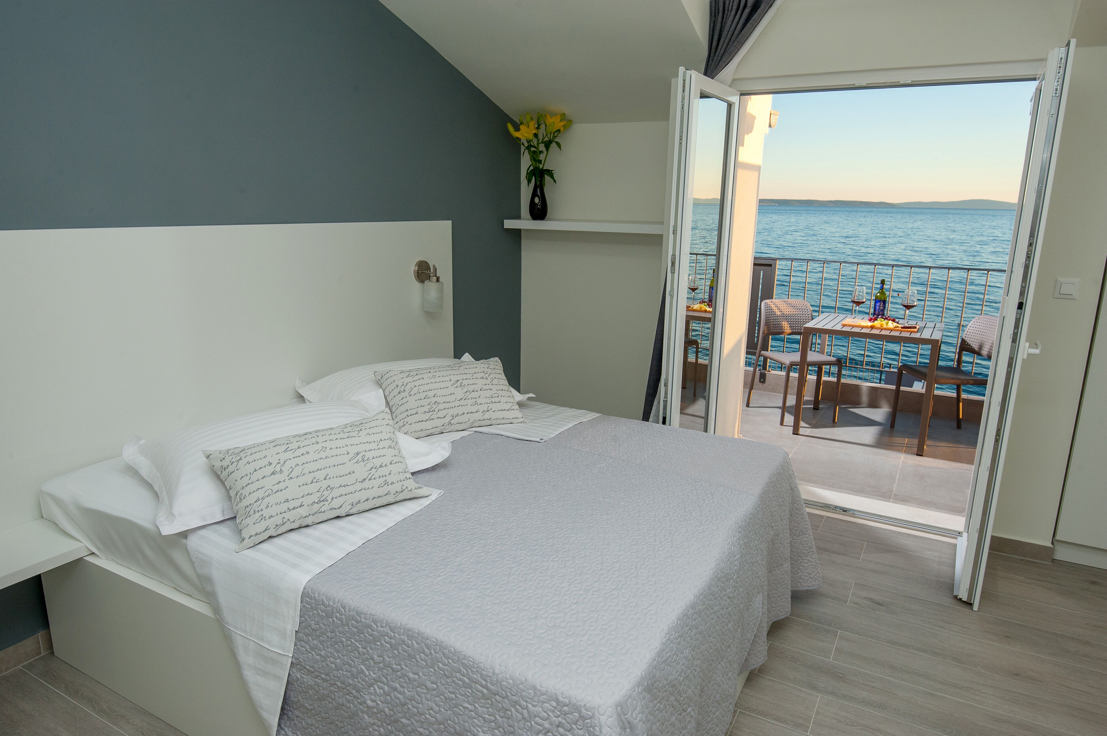 Un dormitorio con cama, mesita de noche, jarra con flores y balcón con mesa y sillas.