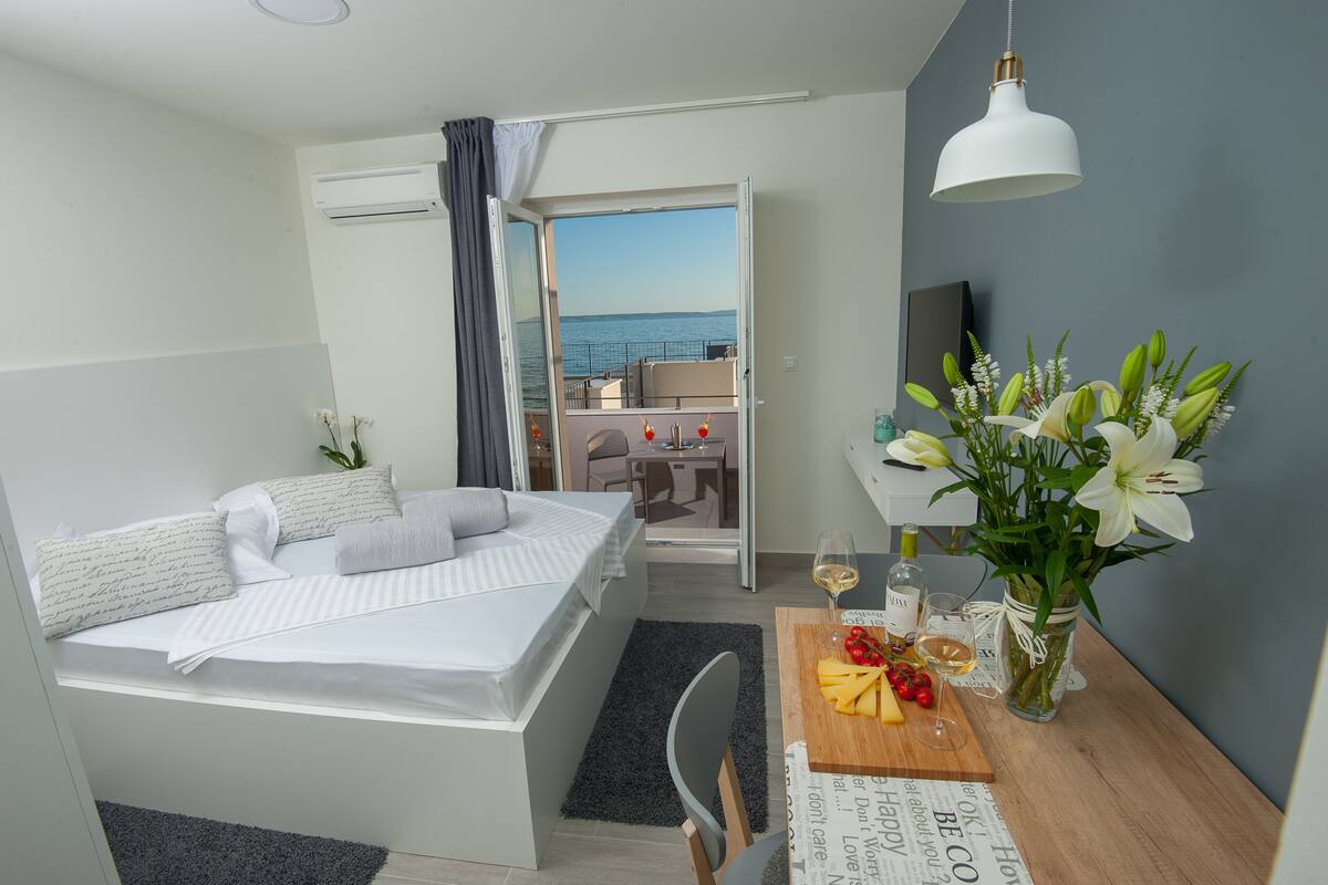 A modern hotel room with a bed, a dining area with a table set for two, a vase of flowers, and a view of the sea through an open door.