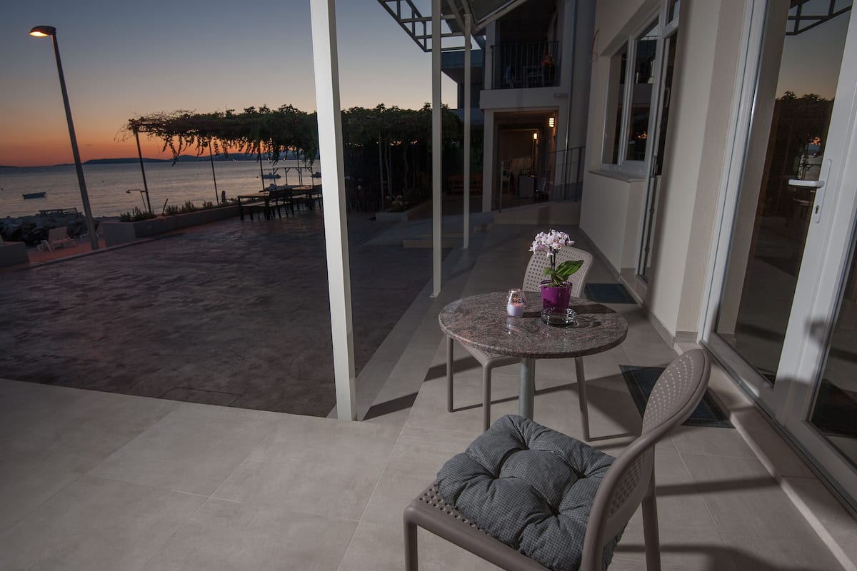 A balcony with a view of the sea at sunset, a round table with a cushion, and a chair.