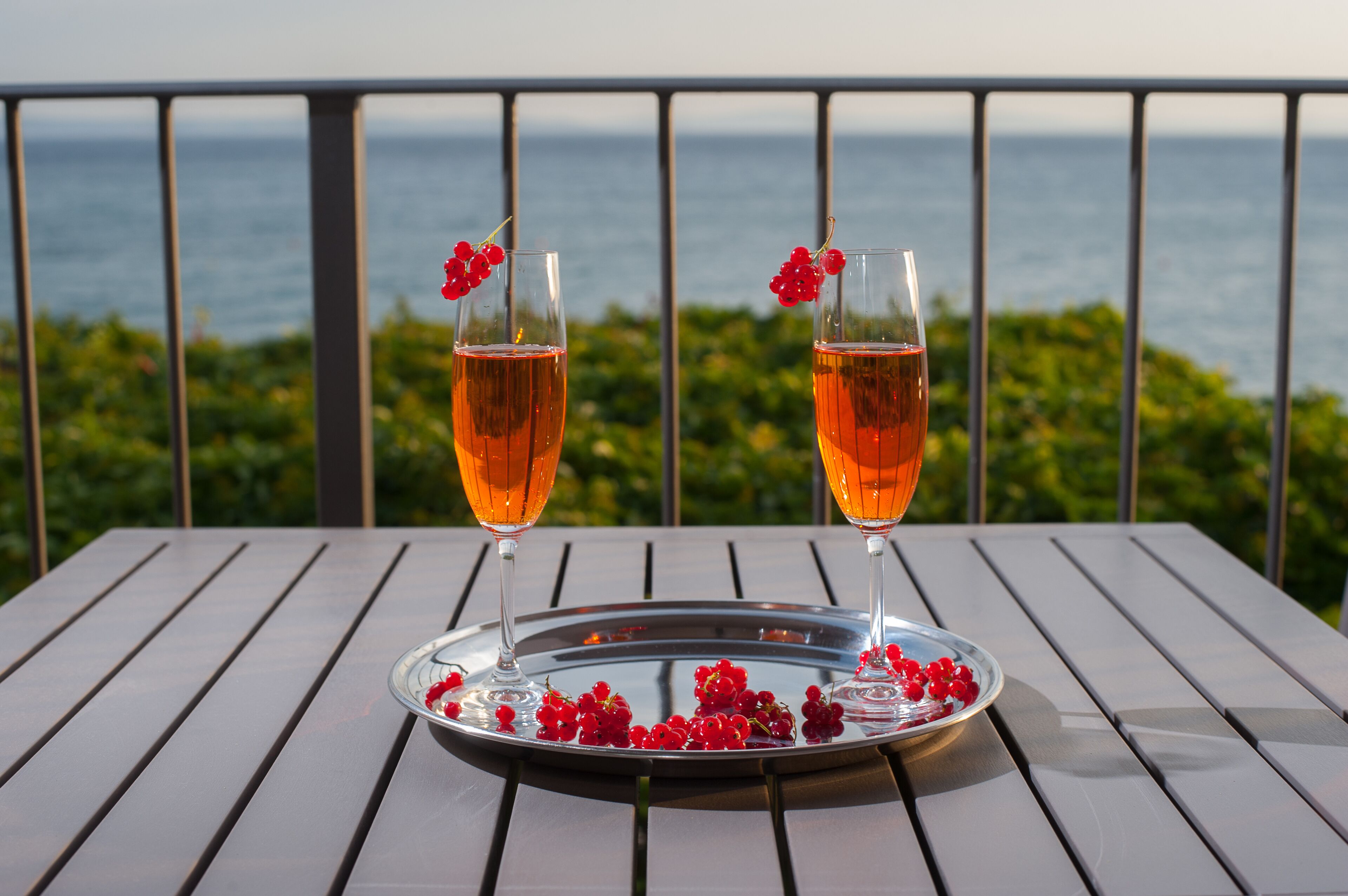 Deux verres d’un liquide ambré, posés sur un plateau en métal avec des baies rouges, sont placés sur une table en bois. On aperçoit la mer et la verdure en arrière-plan.