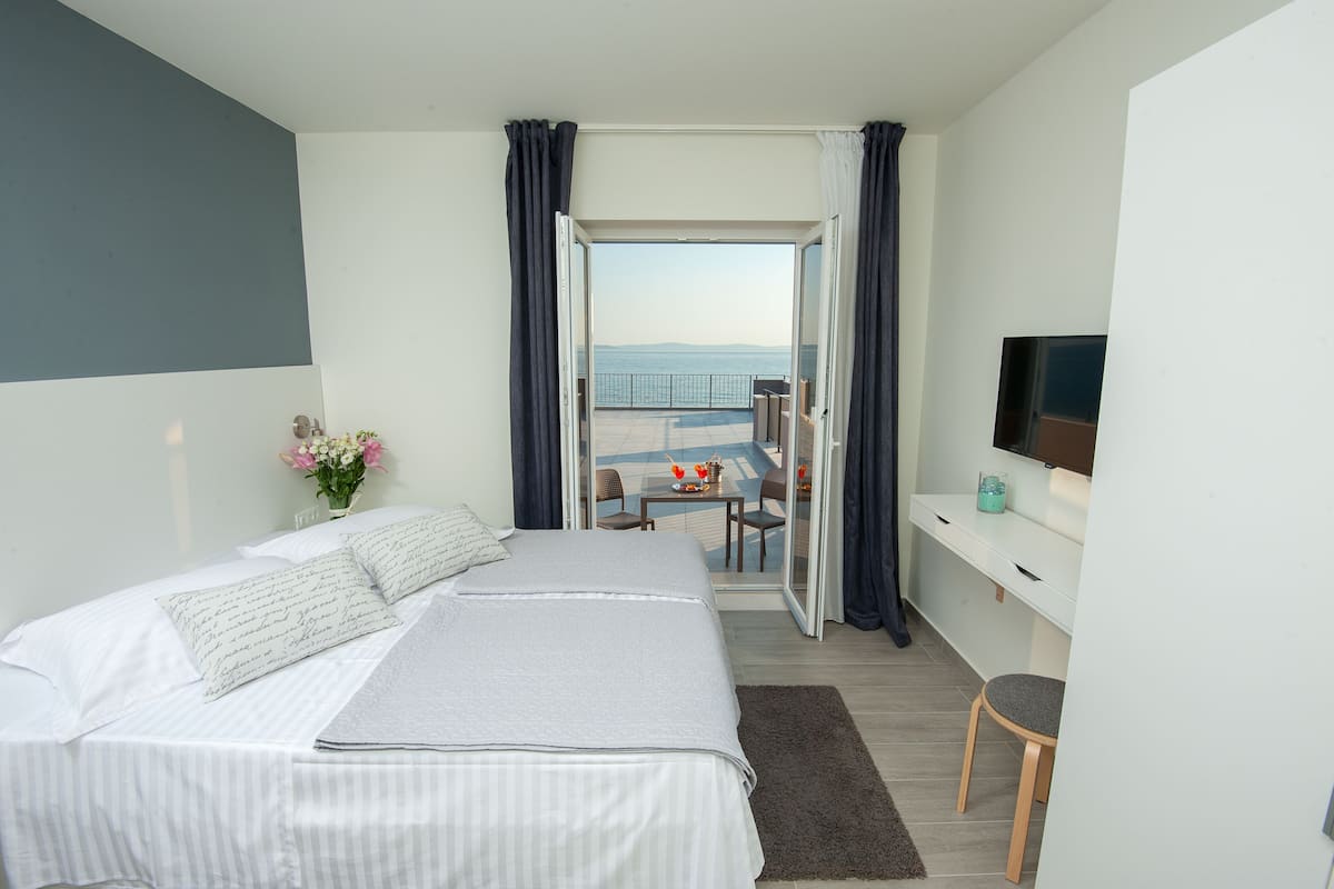 A modern bedroom with a bed, a desk, and a TV. It has a balcony with a table and chairs, and a view of the sea.