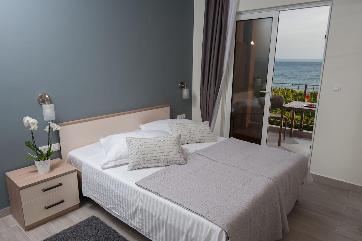 A bedroom with a bed, bedside tables, a potted plant, and a balcony with a view of the sea.