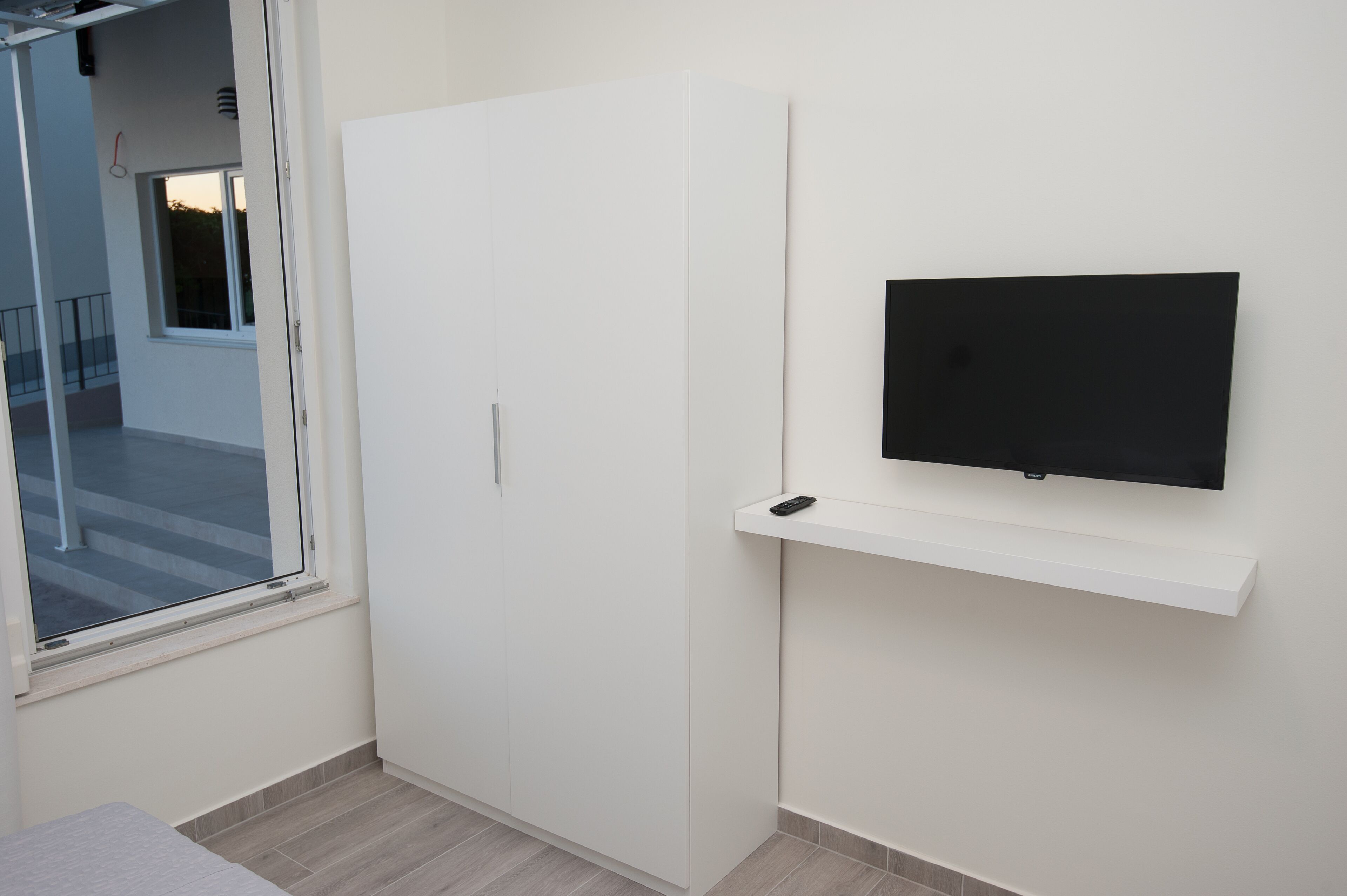 Una stanza con un armadio bianco, una TV a muro e una finestra che dà su una scala e una ringhiera.