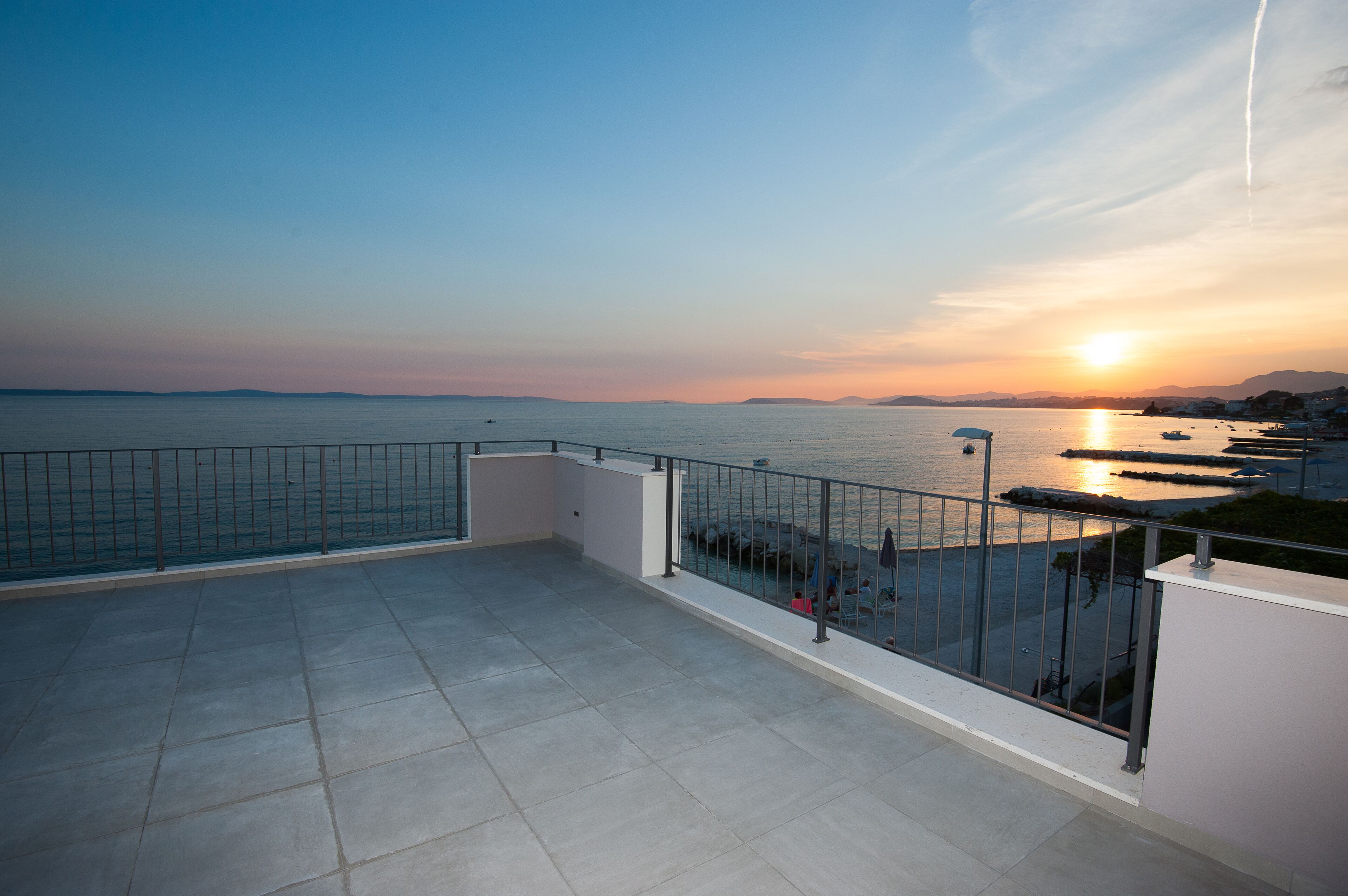 A rooftop terrace with a view of the sea at sunset, featuring a railing and a tiled floor.