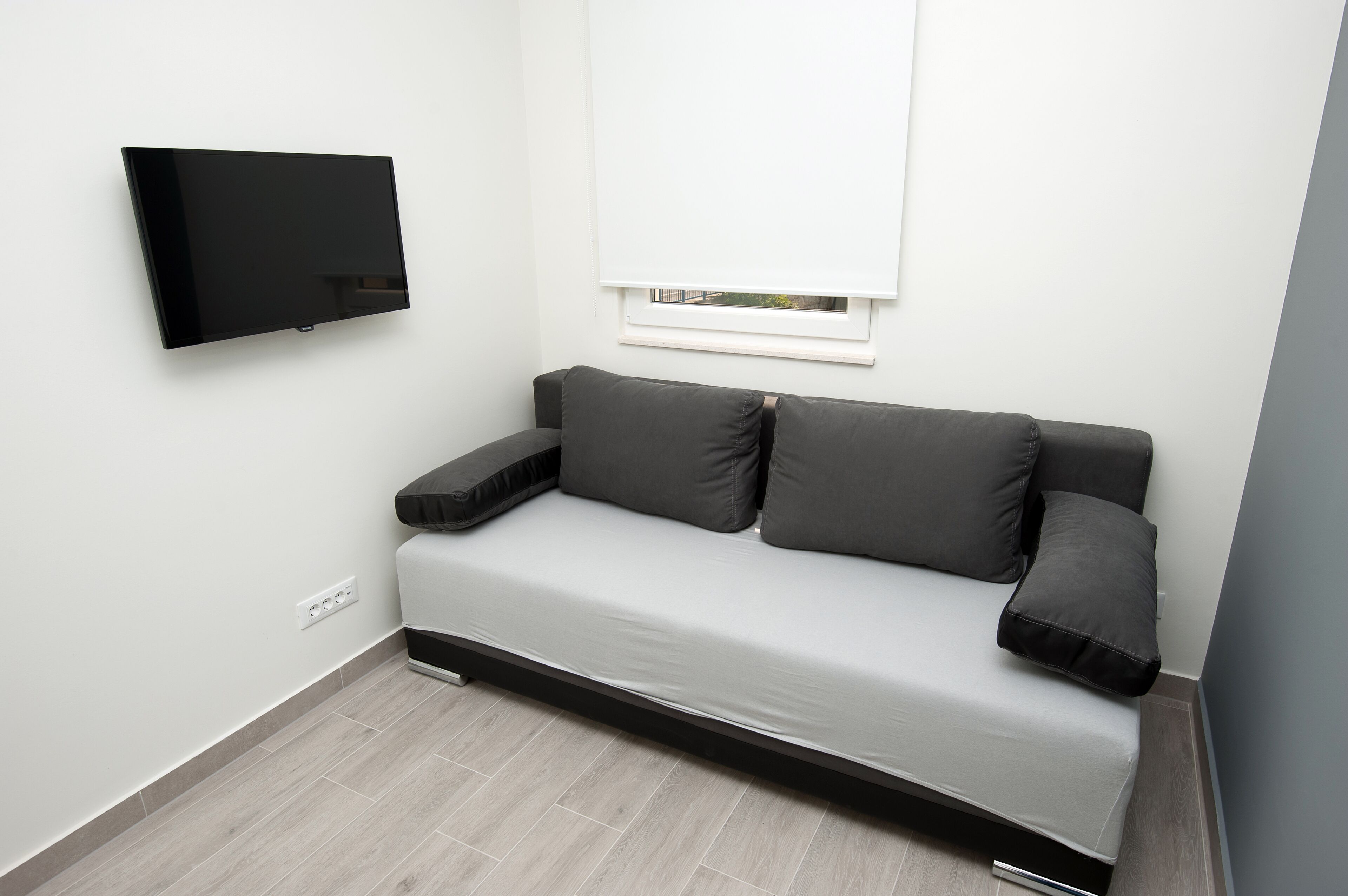 A modern, minimalist room with a grey sofa, a flat-screen TV mounted on the wall, and a window with blinds.