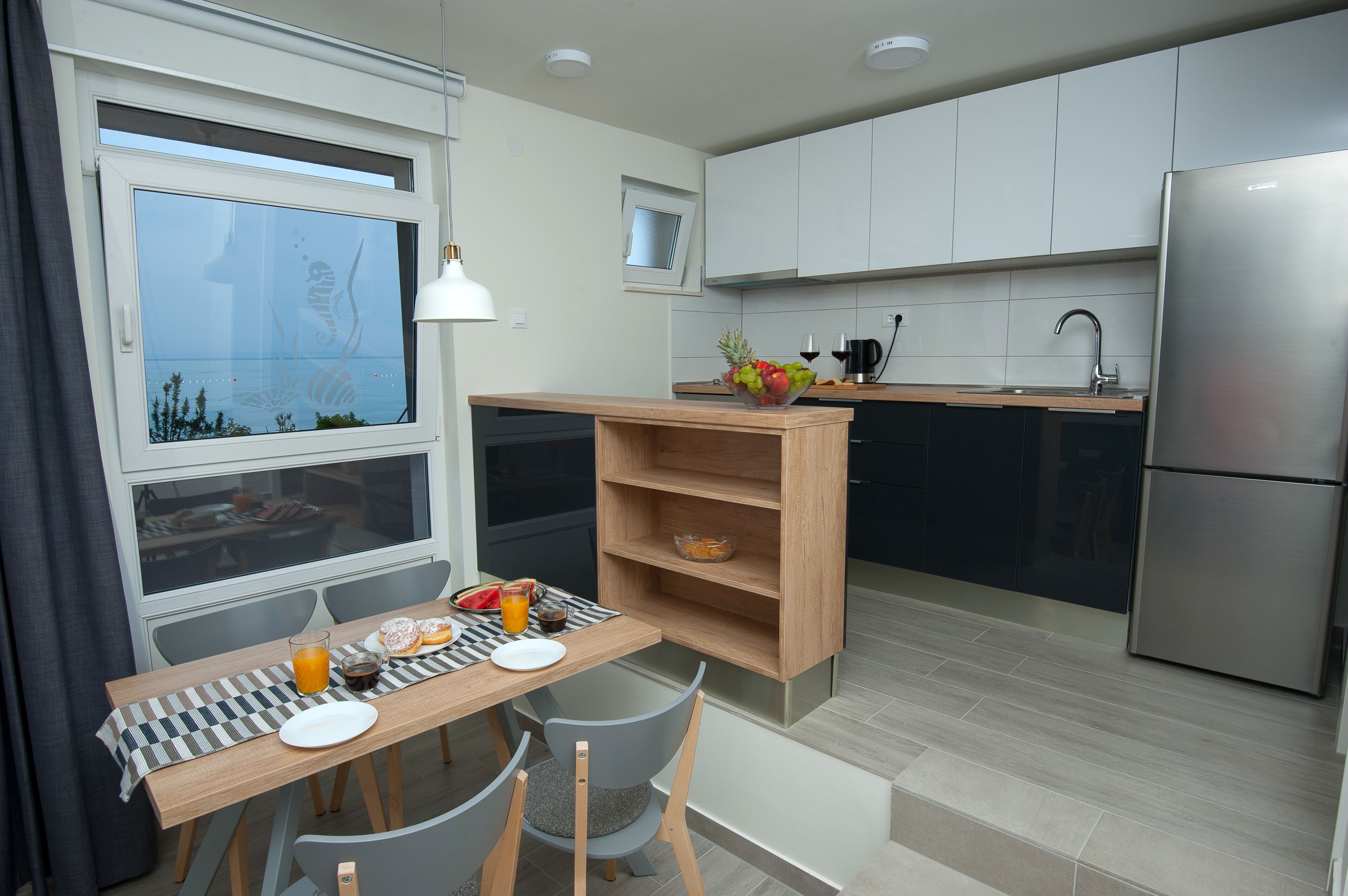 A modern kitchen with a dining area, featuring a wooden table, chairs, a bowl of fruit, and a refrigerator.