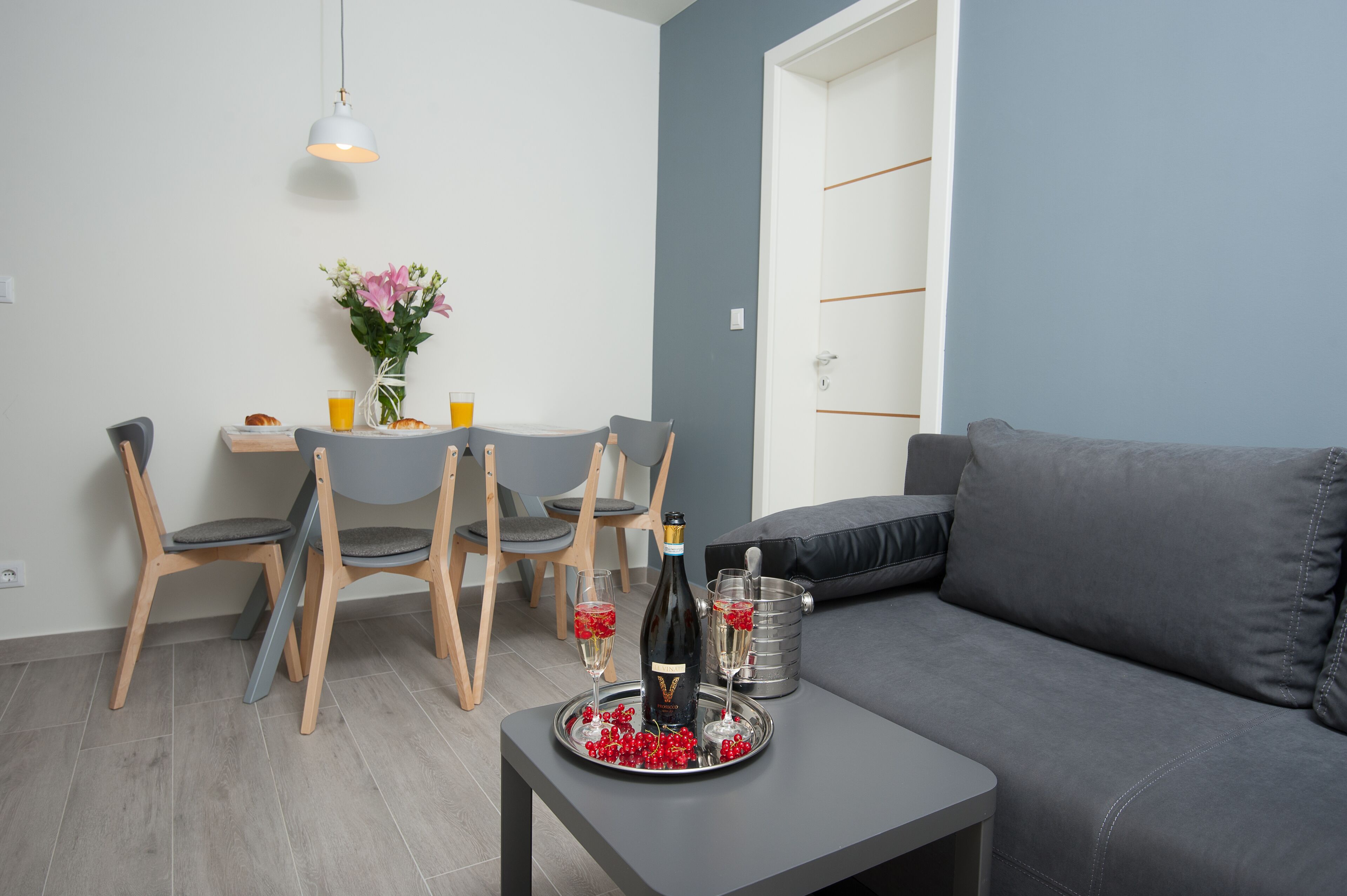 A modern living room with a grey sofa, a dining table with chairs, and a small coffee table with a bottle and glasses.