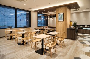 A modern restaurant with wooden tables and chairs, large windows, and a central bar area.