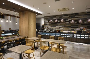 Interior moderno de restaurante con mesas y sillas de madera, una barra con exhibición de alimentos y una pared con frascos de condimentos.