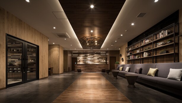 A modern lobby with a wooden reception desk, a dark-colored sofa with cushions, and a large bookshelf filled with books. - Nest Hotel Hakata Station (Fukuoka)