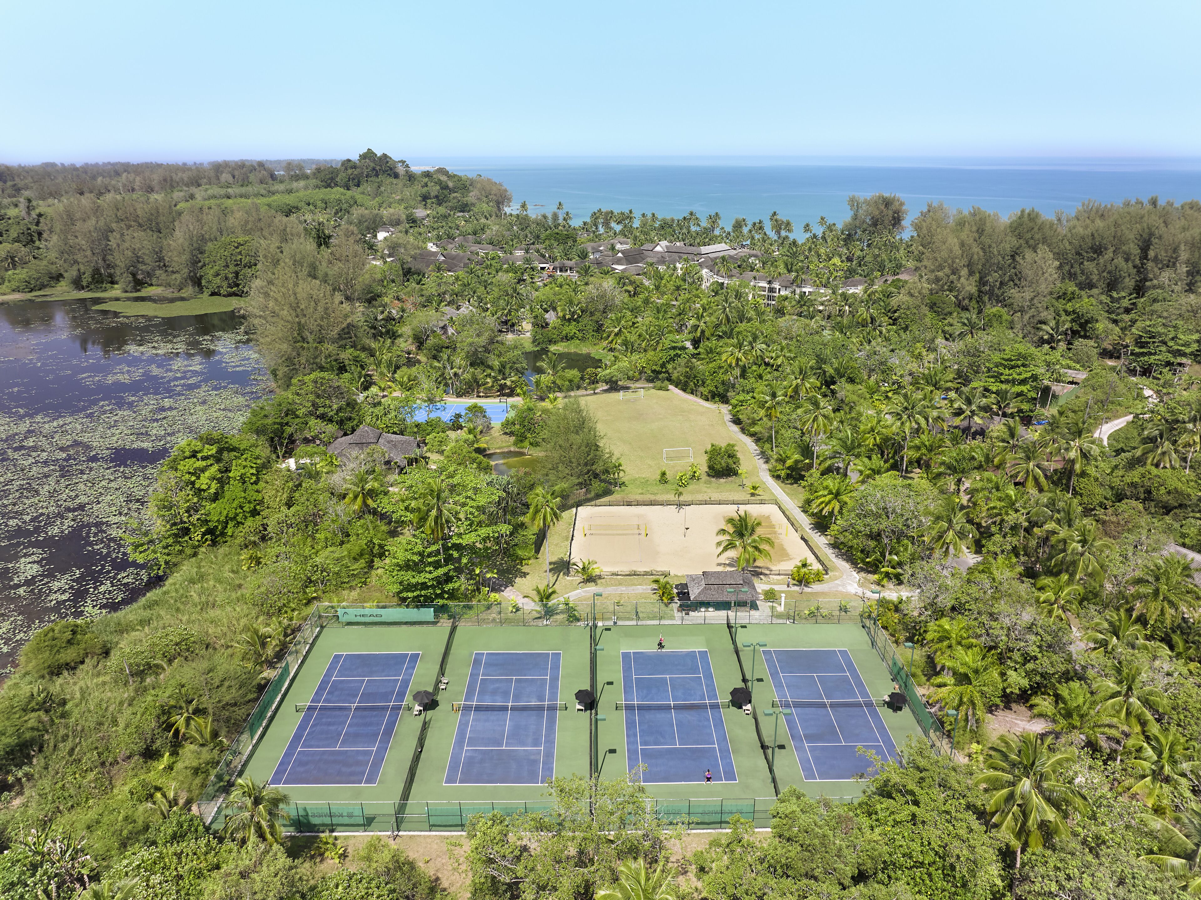 An aerial view of a tennis court complex surrounded by lush greenery and a lake, with a clear view of the ocean in the background.