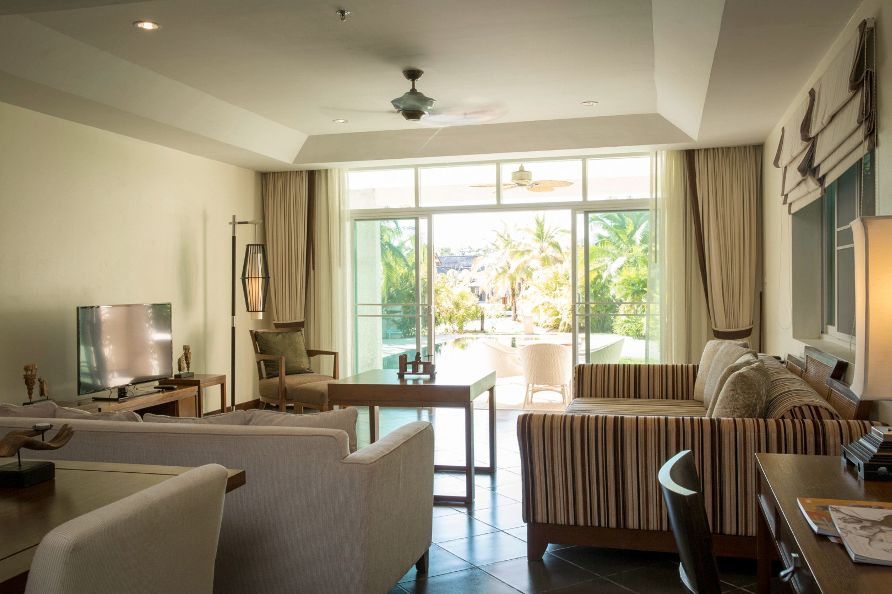 A modern living room with a flat-screen TV, a sofa, a dining table, and a large window offering a view of palm trees.