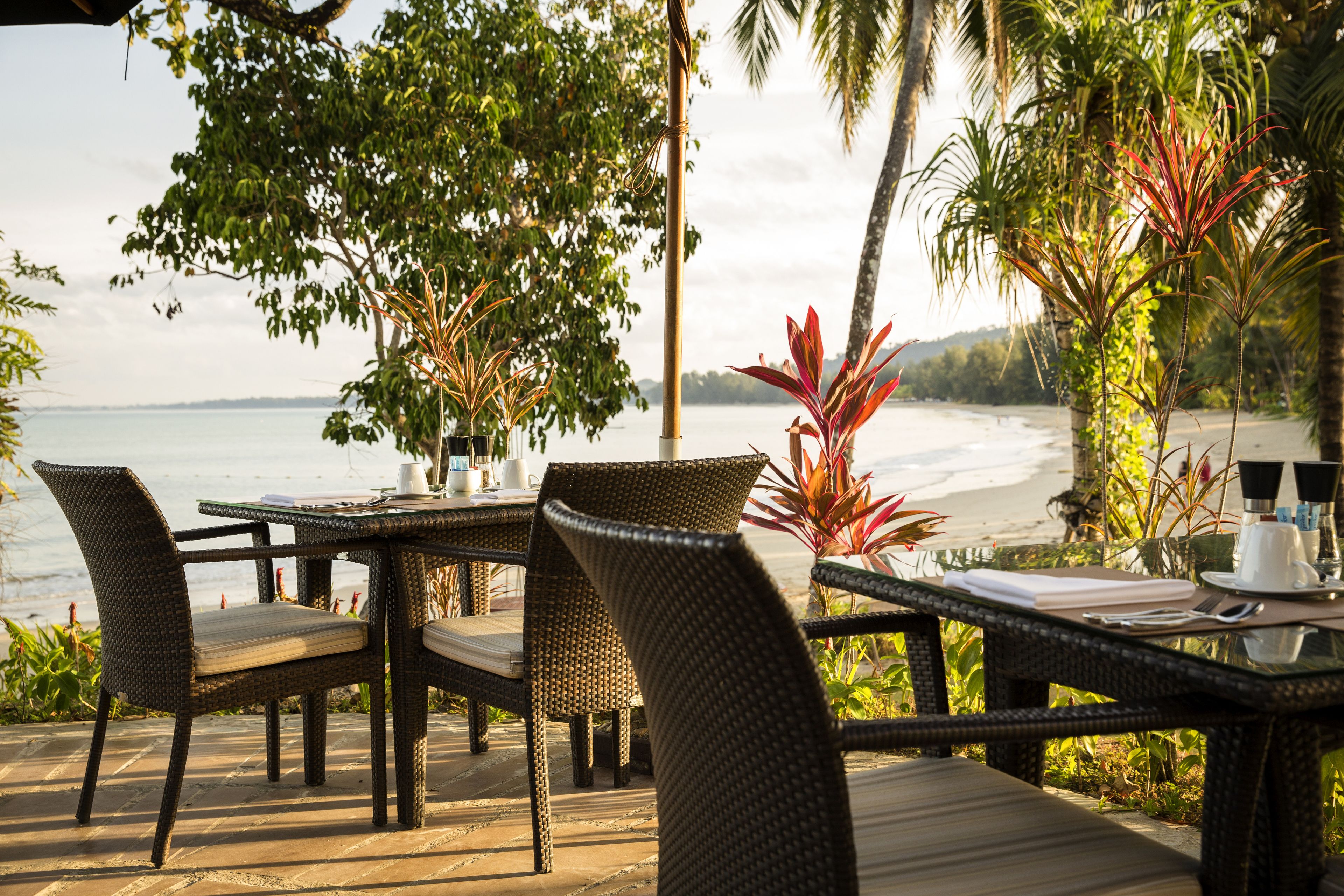 A dining area with wicker furniture, a view of the beach, and tropical plants.