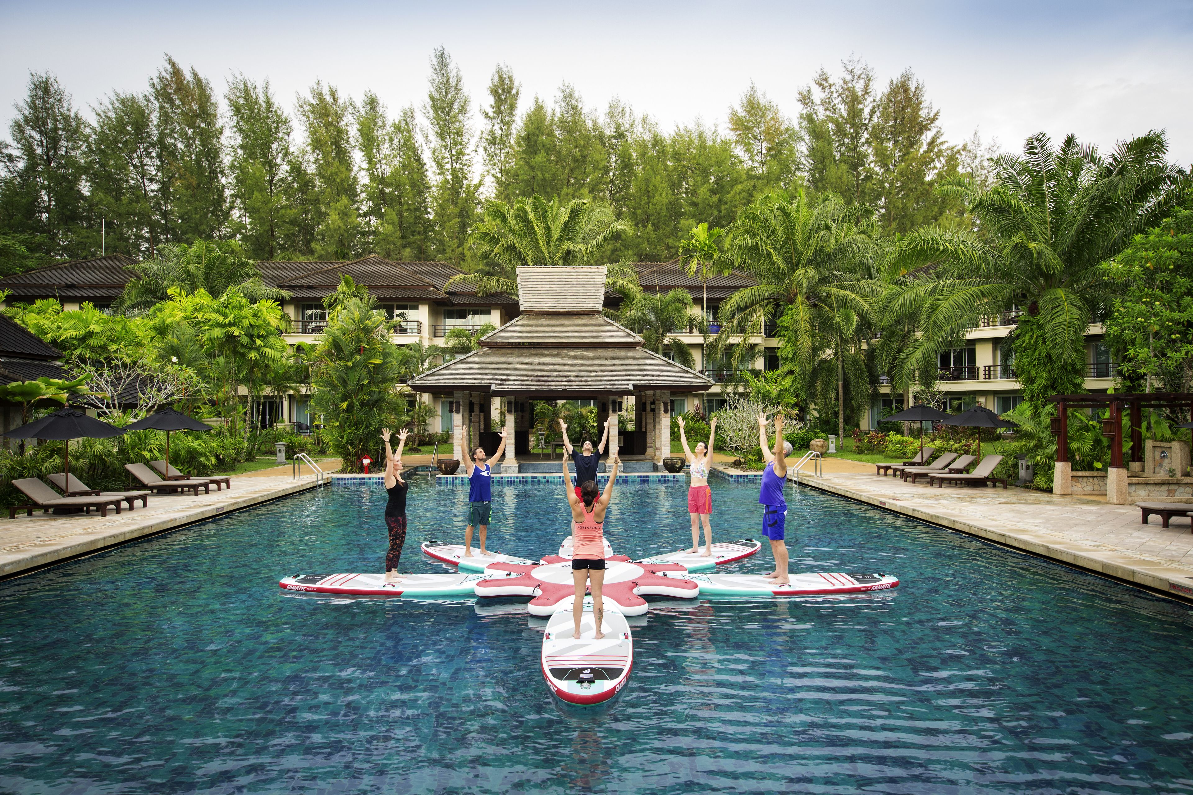 A swimming pool with people on floating platforms, surrounded by palm trees and lounge chairs.