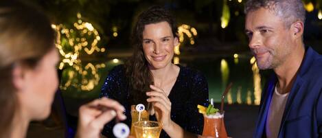 Tres personas sentadas en una mesa al aire libre con bebidas, rodeadas de luces decorativas.