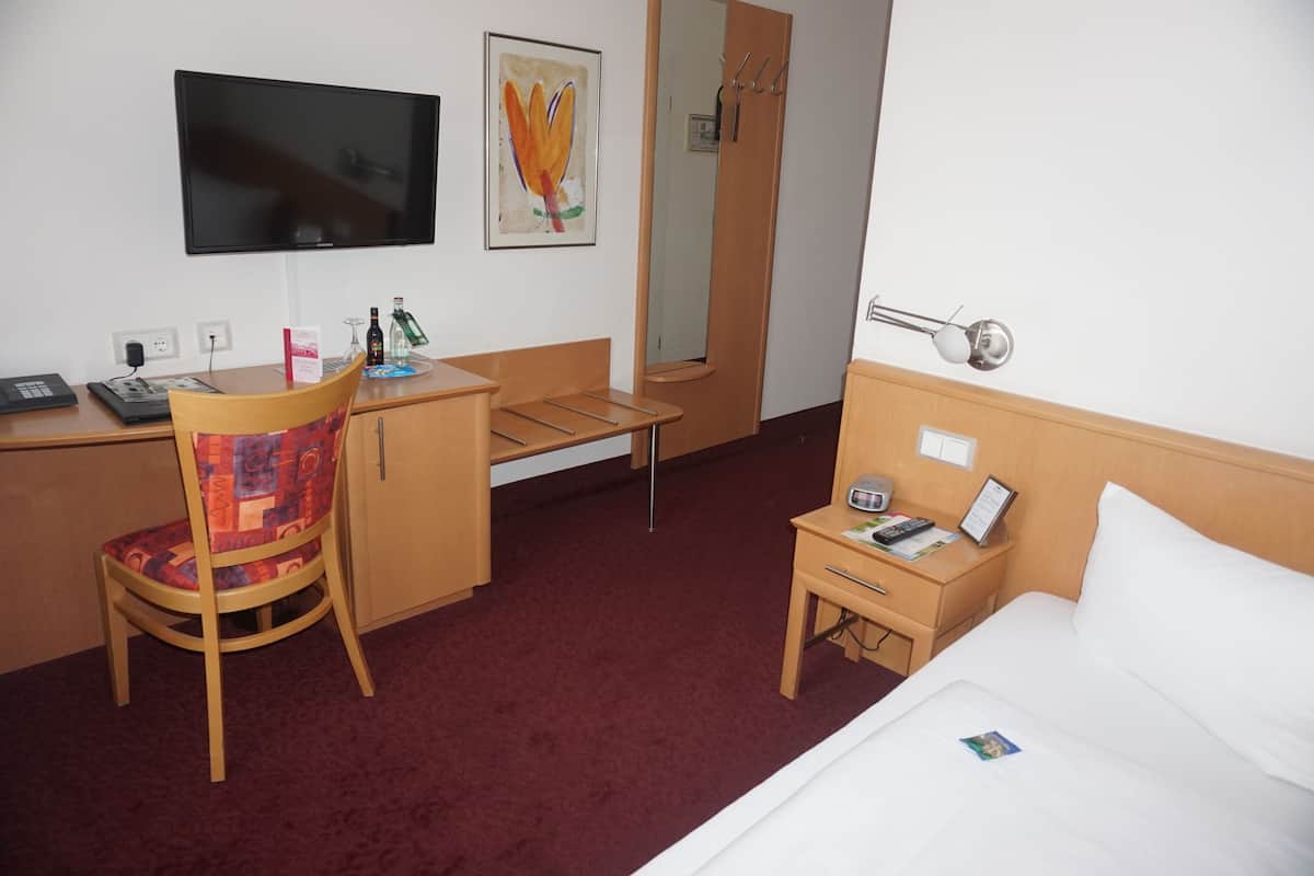Une chambre d’hôtel comprenant un lit, un bureau avec une chaise, une télévision, un miroir et une petite table avec une lampe.