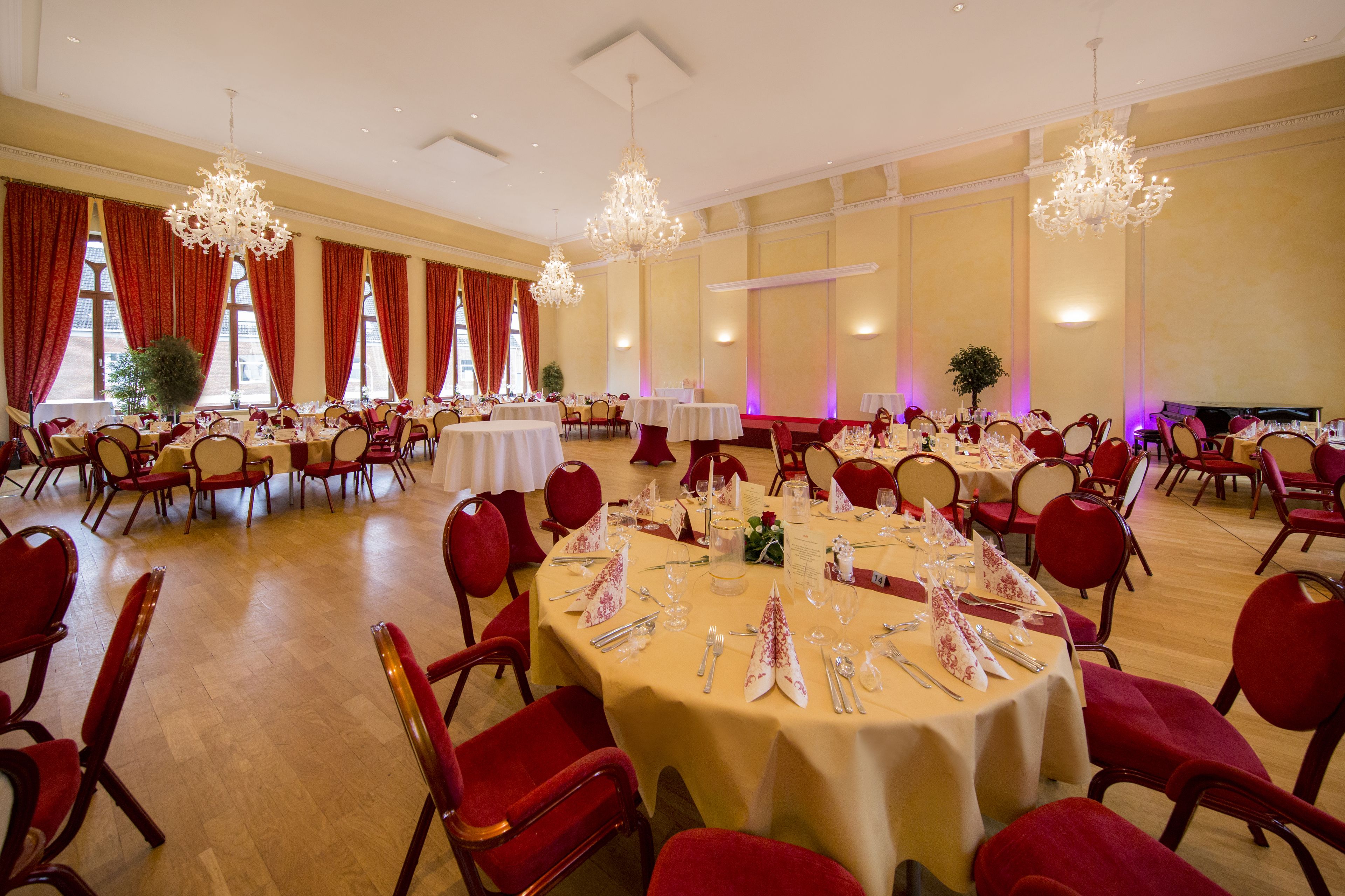 A banquet hall with round tables set for dining, red upholstered chairs, and chandeliers.