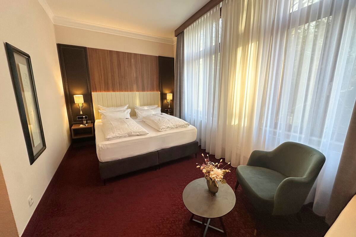 Une chambre d’hôtel avec un grand lit, un fauteuil vert, une petite table ronde ornée d’un vase de fleurs et une grande fenêtre avec des rideaux légers.