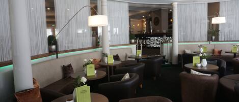 A modern hotel lobby with round tables, comfortable seating, and a bar area in the background.