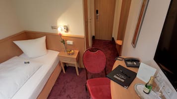 Una habitación de hotel con una cama, una mesita de noche con lámpara, una silla, un escritorio con teléfono y libreta, y una puerta que da a otra habitación.