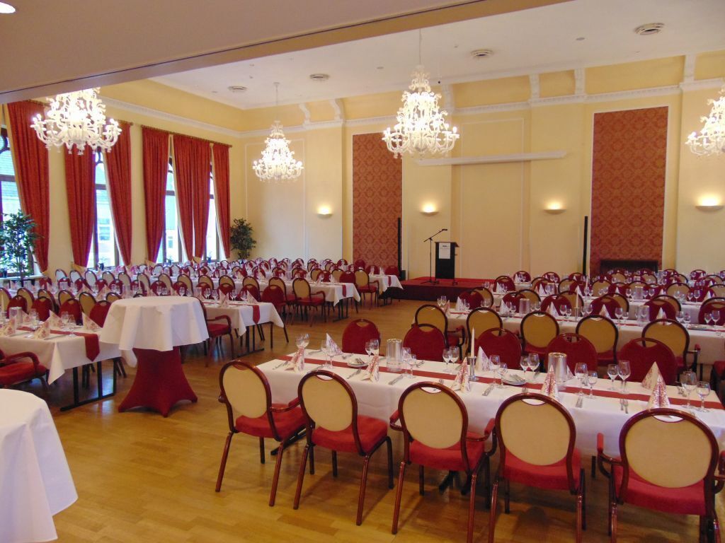 Un salón de banquetes con filas de mesas dispuestas para comer, candelabros y un escenario para presentaciones.