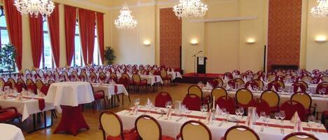 Un salón de banquetes con filas de mesas dispuestas para comer, candelabros y un escenario para presentaciones.