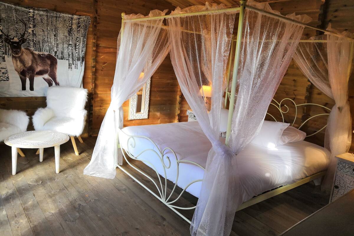 A rustic wooden cabin with a four-poster canopy bed, a white furry chair, and a small round table.