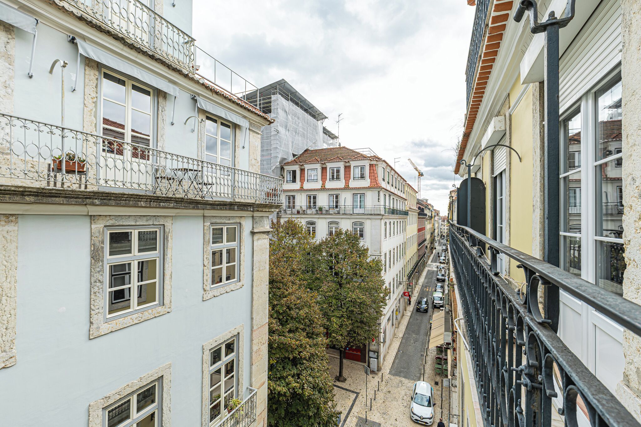A narrow street with buildings on either side, featuring balconies with metal railings and potted plants.