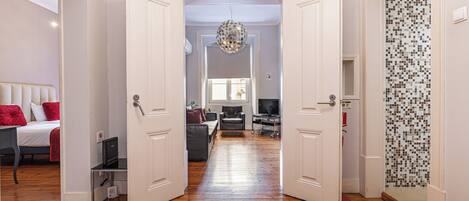A hallway leading to a bedroom with a bed, a desk, and a chair. There is a door on the right leading to another room with a tiled wall.
